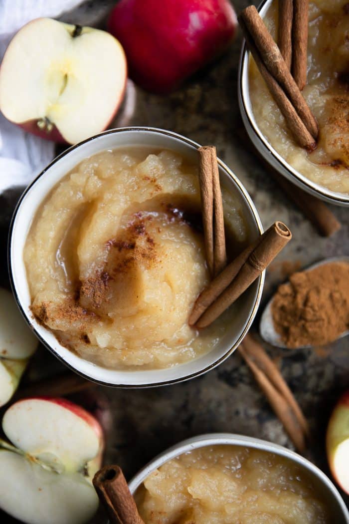 Overhead image of three small bowls filled with homemade applesauce garnished with ground cinnamon and two whole cinnamon sticks.