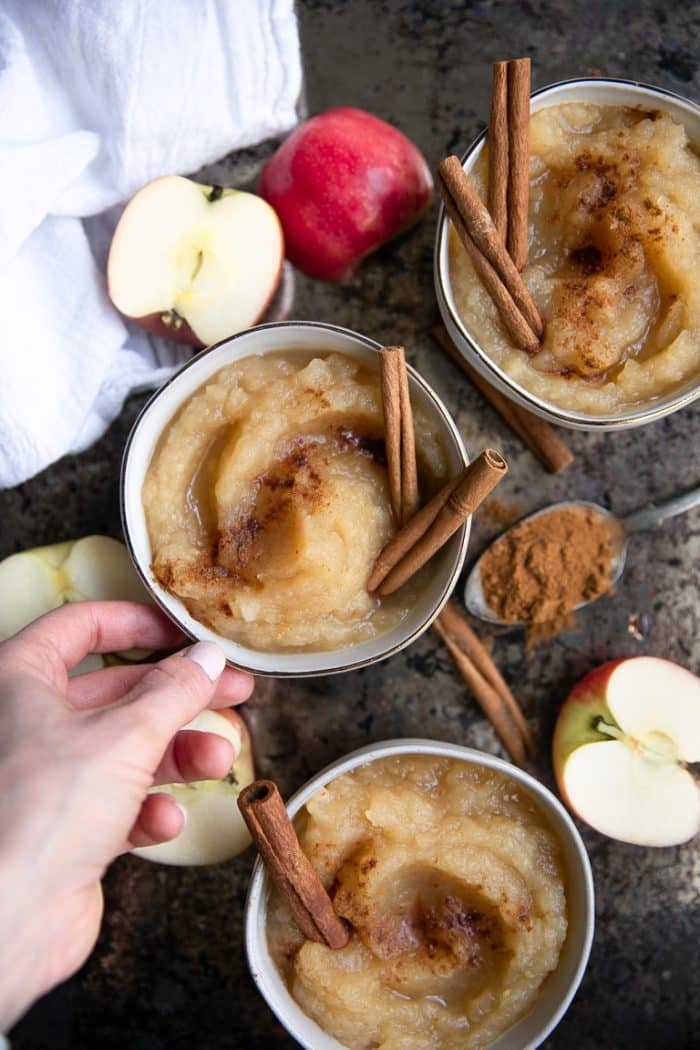 Overhead image of three small bowls filled with homemade applesauce garnished with ground cinnamon and two whole cinnamon sticks with halved apples in the background.
