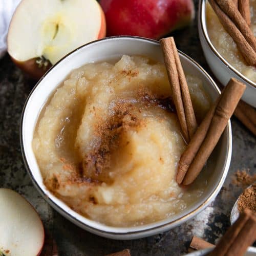 Close-up image of a bowl filled with homemade applesauce made with pink lady apples and topped with ground cinnamon and whole cinnamon sticks.