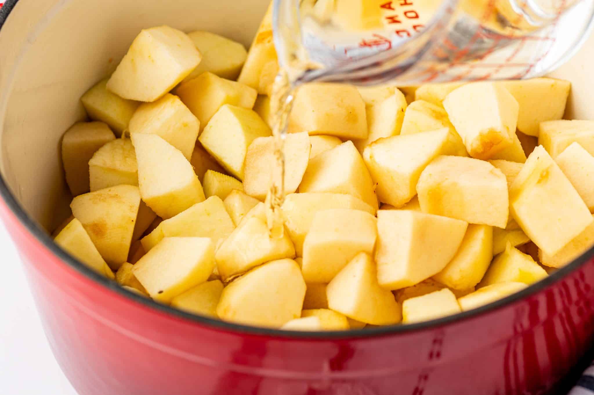 stream of water pours from a clear glass measuring cup with red markings onto peeled apple chunks in a red enamel pot with a cream-colored interior. The pale yellow apple pieces remain uncovered by the small amount of liquid.