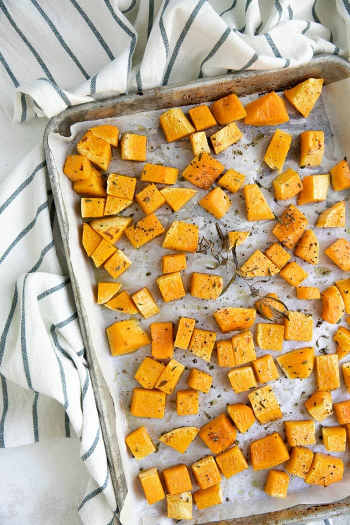 Roasted cubes of butternut squash on a large baking sheet in a single layer seasoned with fresh herbs.