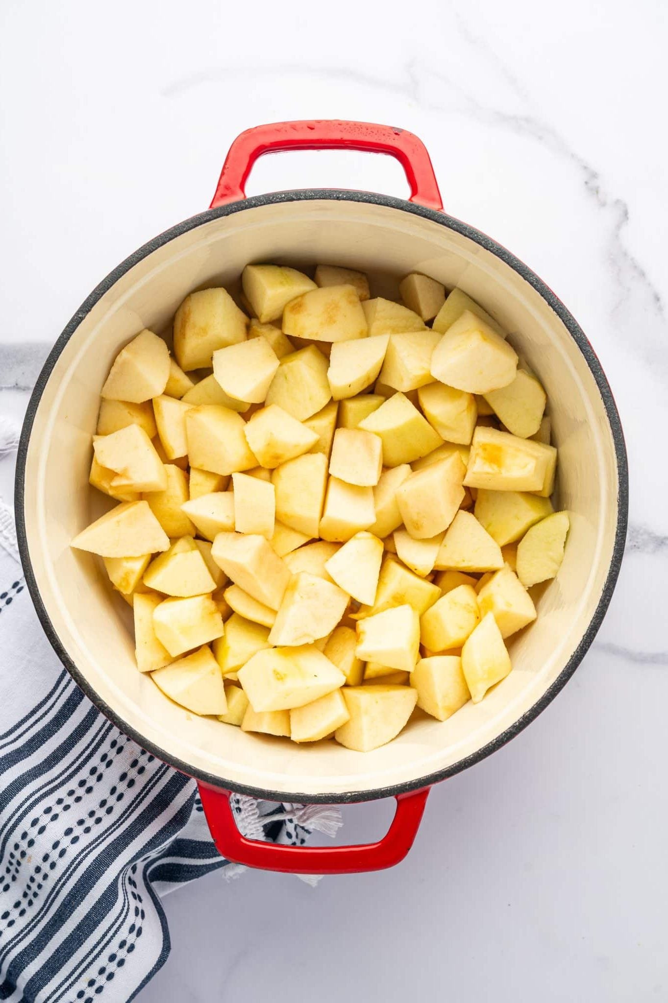 Peeled and cored pale yellow apple chunks fill a cream-colored enamel pot with red handles. The apples are cut into irregular wedges and smaller pieces, with no seasonings visible on top. A navy-and-white patterned towel rests partly beneath the pot on a white marble surface.