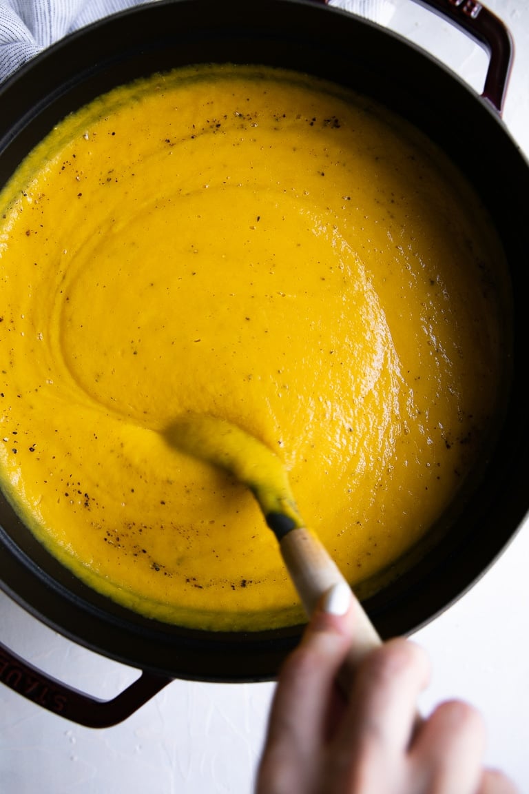 Hand holding a wooden spoon stirring a large cast iron dutch oven filled with creamy butternut squash soup.