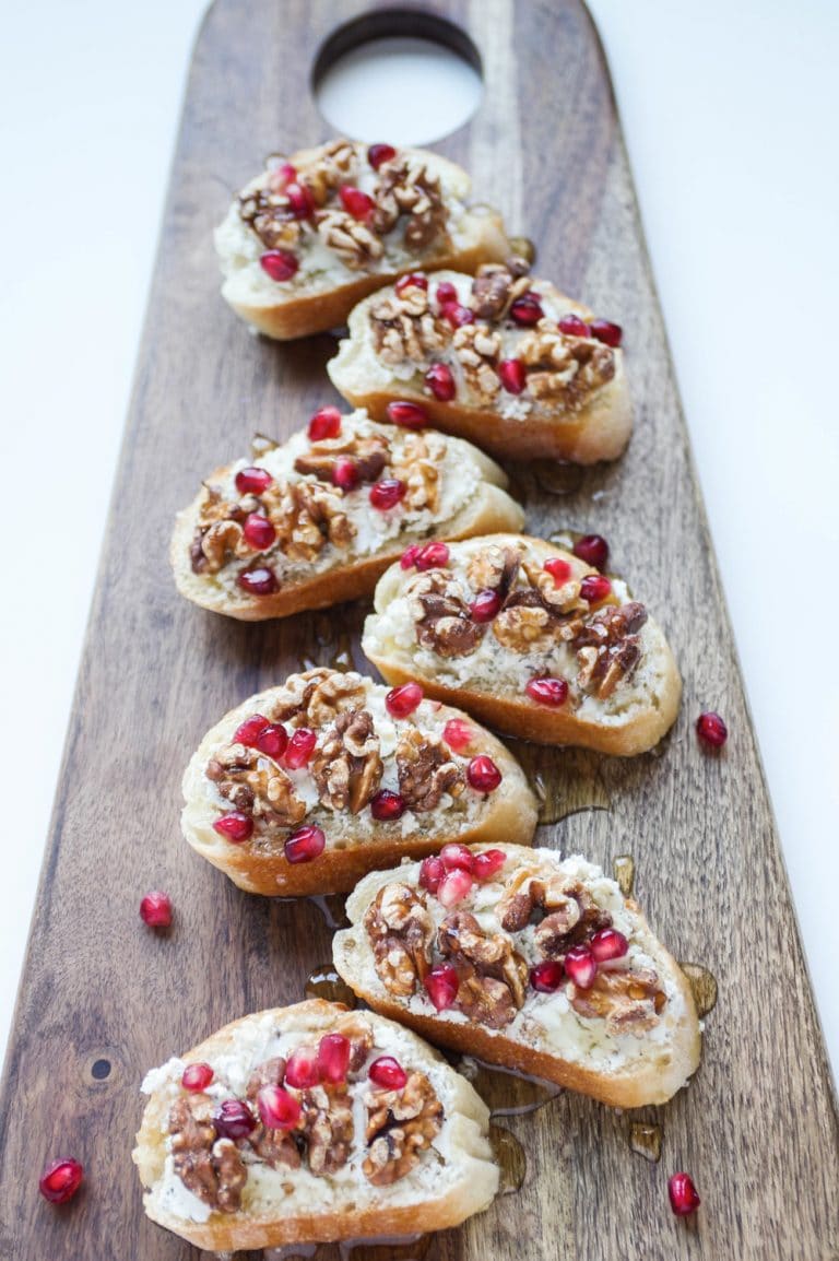 Large serving board with toasted crostini topped with goat cheese, walnuts, pomegranates, and honey.