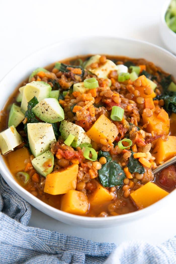 White bowl filled with delicious vegetarian lentil stew filled with chunks of butternut squash, kale, and topped with avocado.