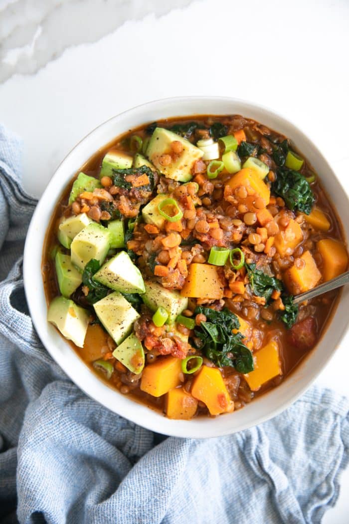 White bowl filled with delicious vegetarian lentil stew filled with chunks of butternut squash, kale, and topped with avocado.