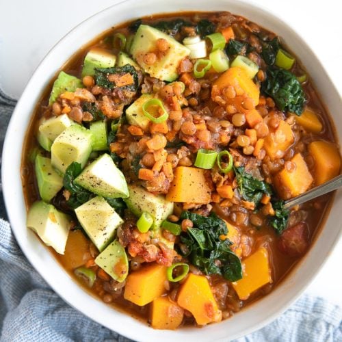 White bowl filled with delicious vegetarian lentil stew filled with chunks of butternut squash, kale, and topped with avocado.