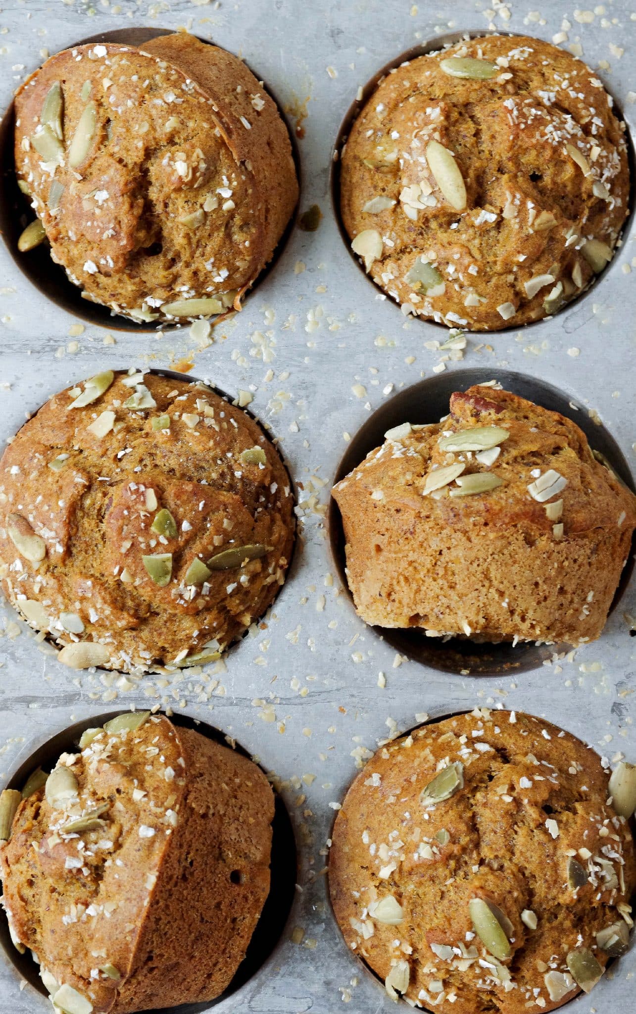 Overhead close-up of six golden-brown pumpkin muffins sitting in a silver muffin tin. The muffins have a domed top and are garnished with green pumpkin seeds and a light dusting of oats, with a few crumbs scattered around the tin.