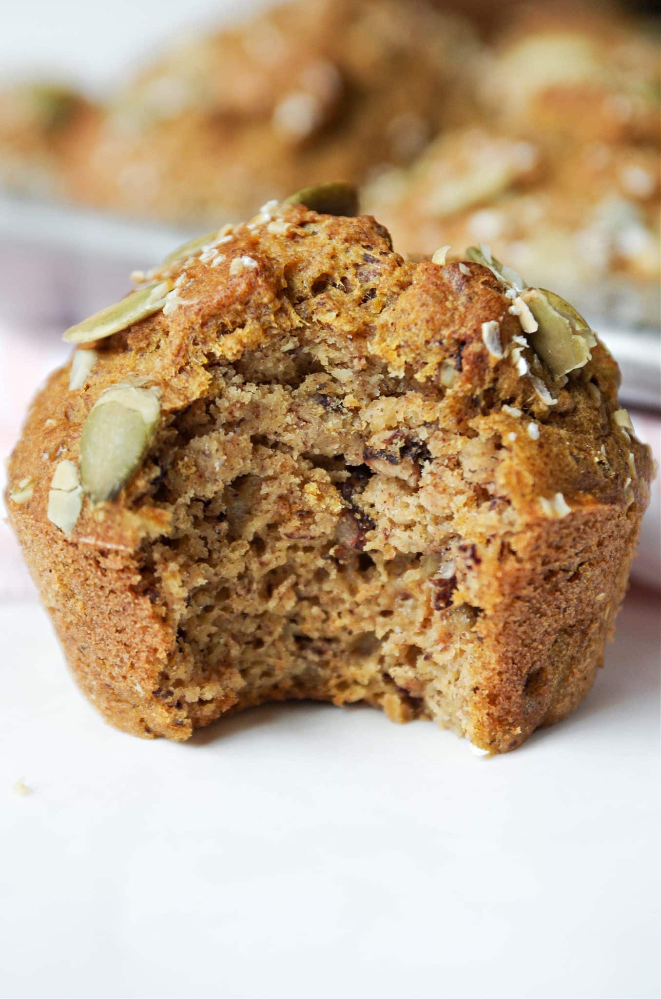A close-up of a pumpkin muffin with a large bite taken out, revealing a tender, moist crumb speckled with seeds. The muffin top is golden brown with pumpkin seeds on top, and the other muffins are softly blurred in the background.