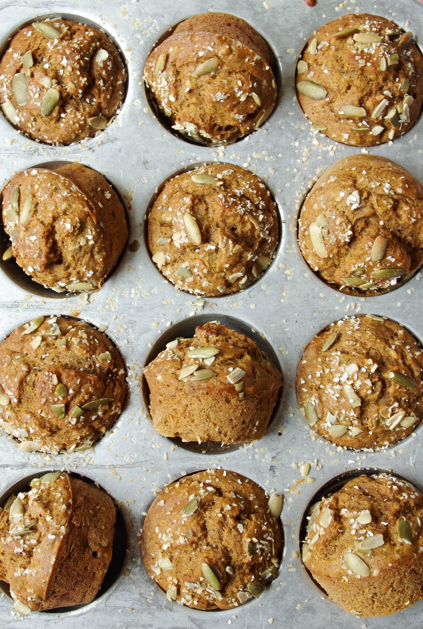 Overhead view of a full muffin tin holding twelve golden-brown pumpkin muffins. Each muffin has a rounded, bakery-style top sprinkled with pumpkin seeds and oats, with light crumbs and seeds scattered on the tin's surface.