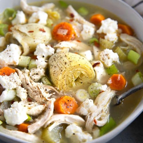 White bowl filled with lemon and artichoke chicken soup and garnished with crumbled feta cheese and red chili flakes.