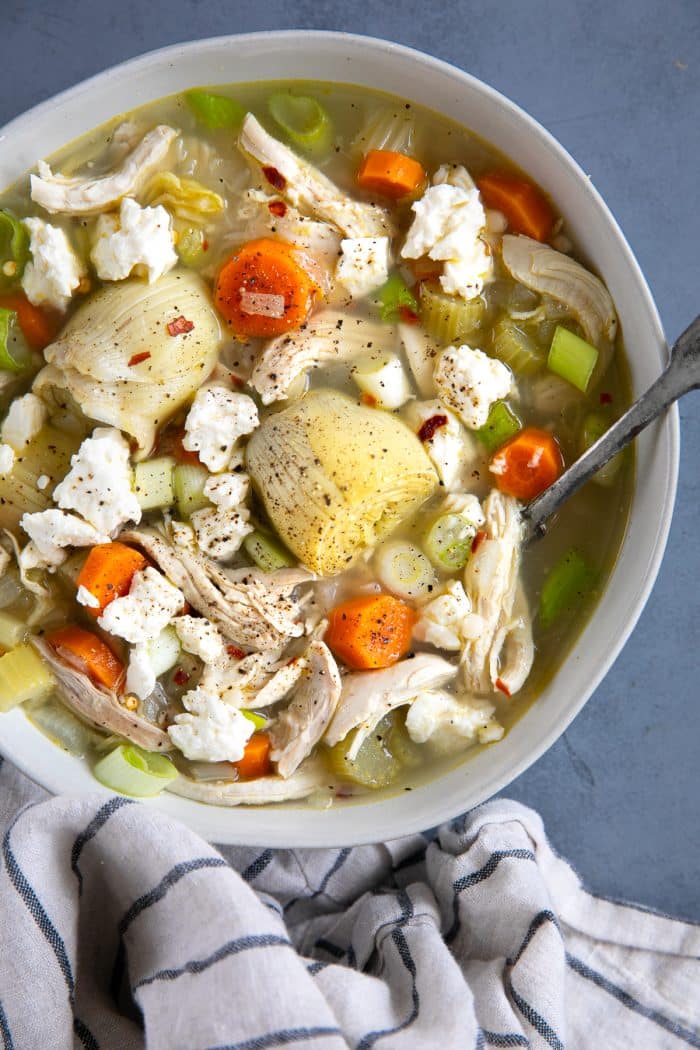 White bowl filled with lemon and artichoke chicken soup and garnished with crumbled feta cheese and red chili flakes.