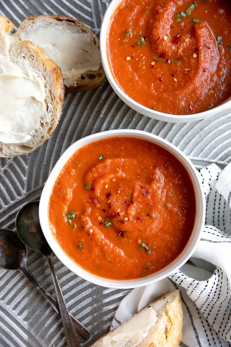 Image of two white bowls filled with homemade tomato basil soup garnished with red chili flakes and fresh basil and served with a side of buttery bread.