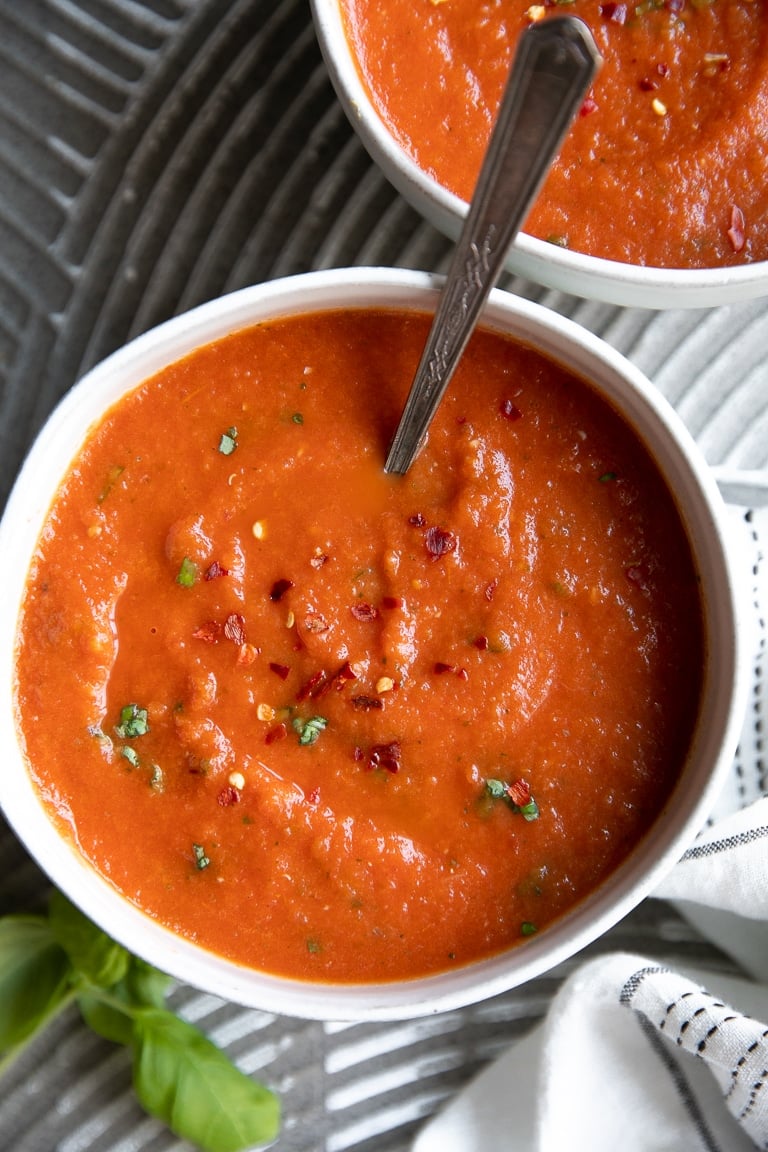 Image of two white bowls filled with homemade tomato basil soup garnished with red chili flakes and fresh basil.
