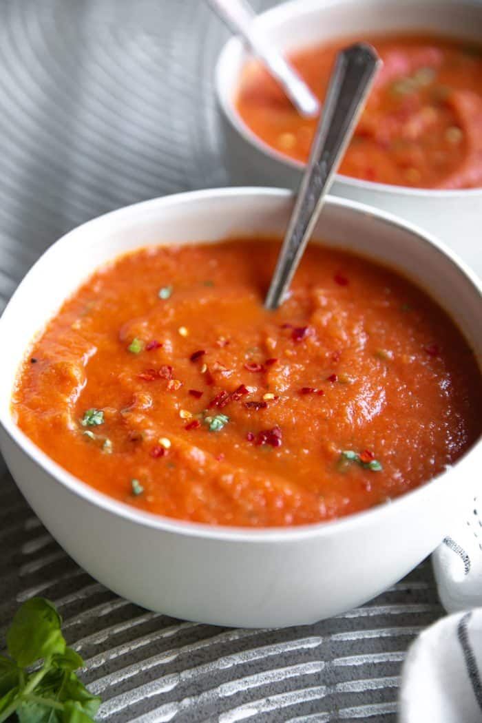 Image of a white bowl filled with cooked and blended homemade tomato basil soup garnished with red chili flakes and fresh basil.