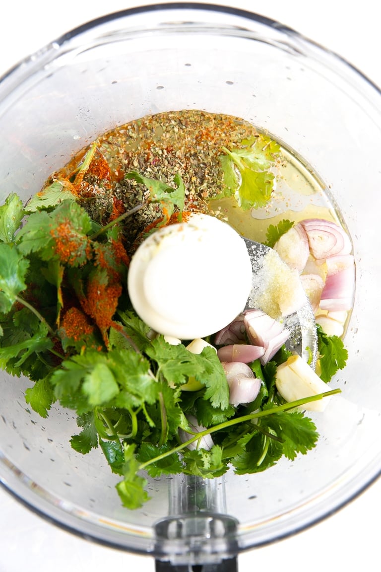Large food processor filled with fresh lime juice, garlic, fresh cilantro leaves, shallots, oregano, salt, black pepper, paprika, olive oil, and red pepper flakes.