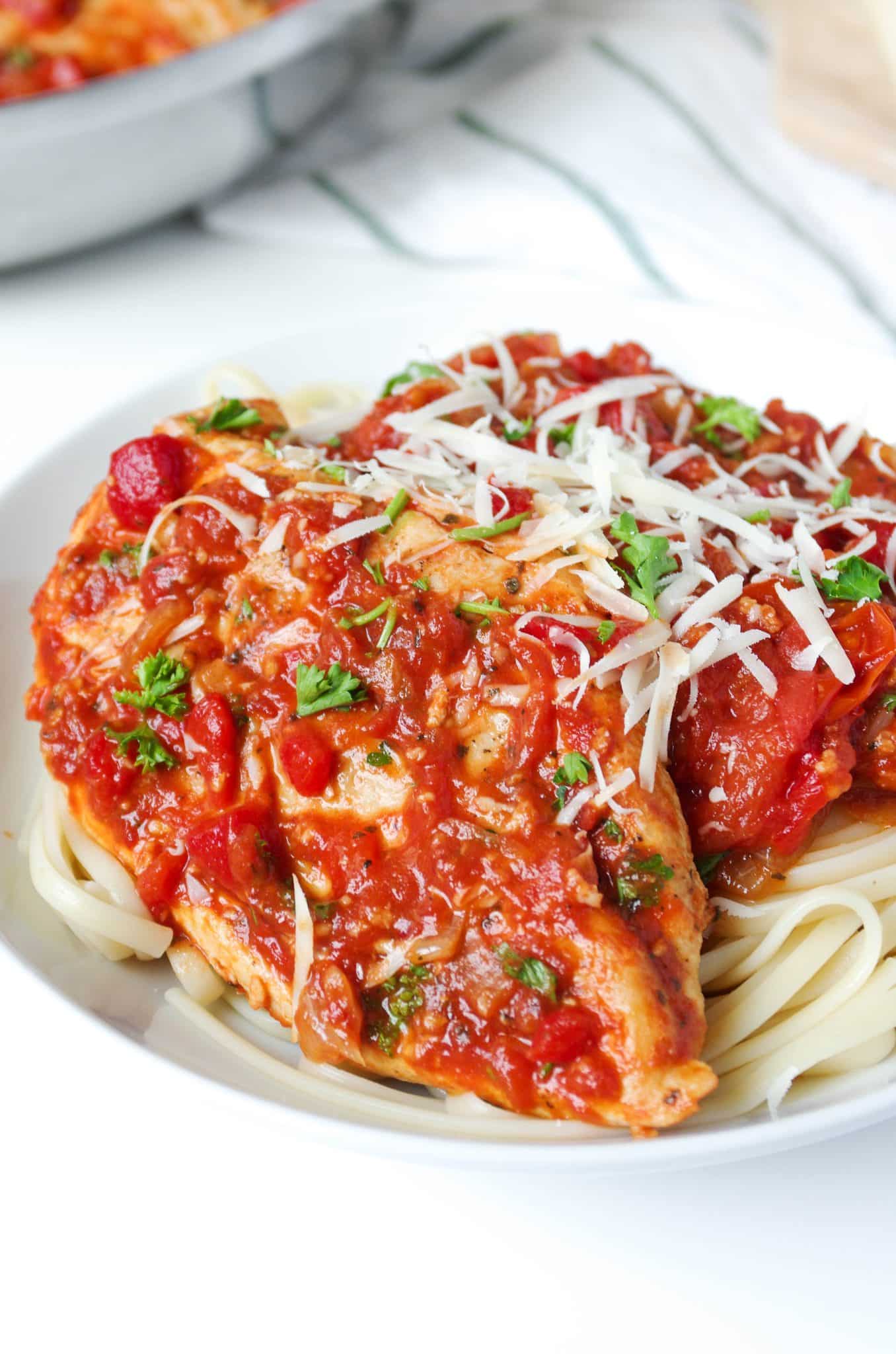 Large white plate filled with linguine paste topped with roasted red pepper and tomato pasta sauce and whole chicken breast garnished with shredded parmesan cheese.