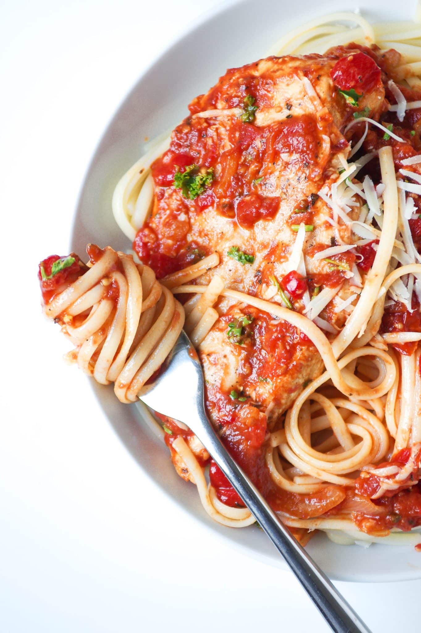Large white plate filled with linguine paste topped with roasted red pepper and tomato pasta sauce and whole chicken breast garnished with shredded parmesan cheese.