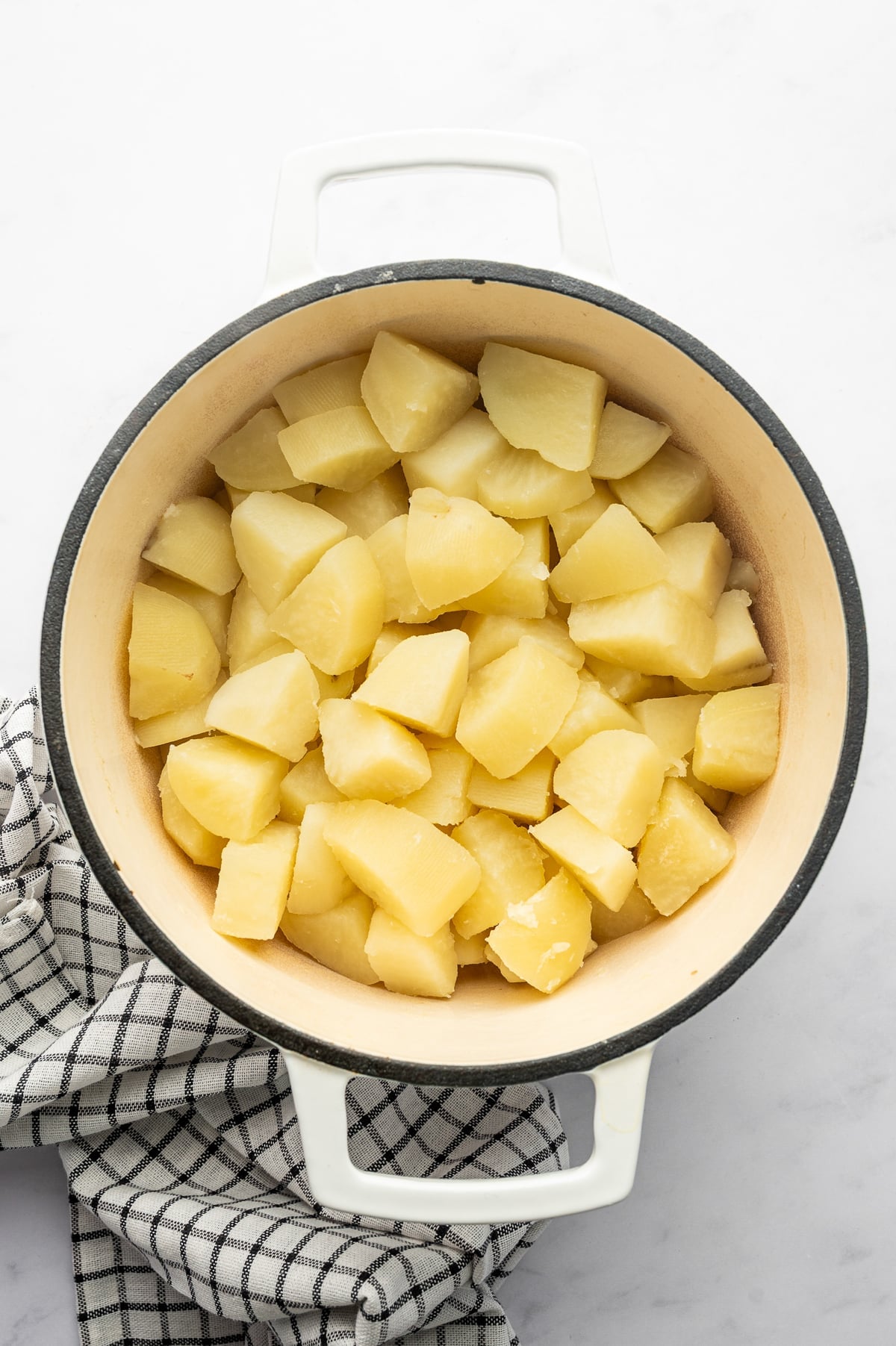 Overhead view of peeled and boiled Russet potato chunks after draining in a white enameled Dutch oven with a cream-colored interior and dark rim, set on a light gray surface beside a black-and-white checkered kitchen towel. No text overlay or garnish is shown.