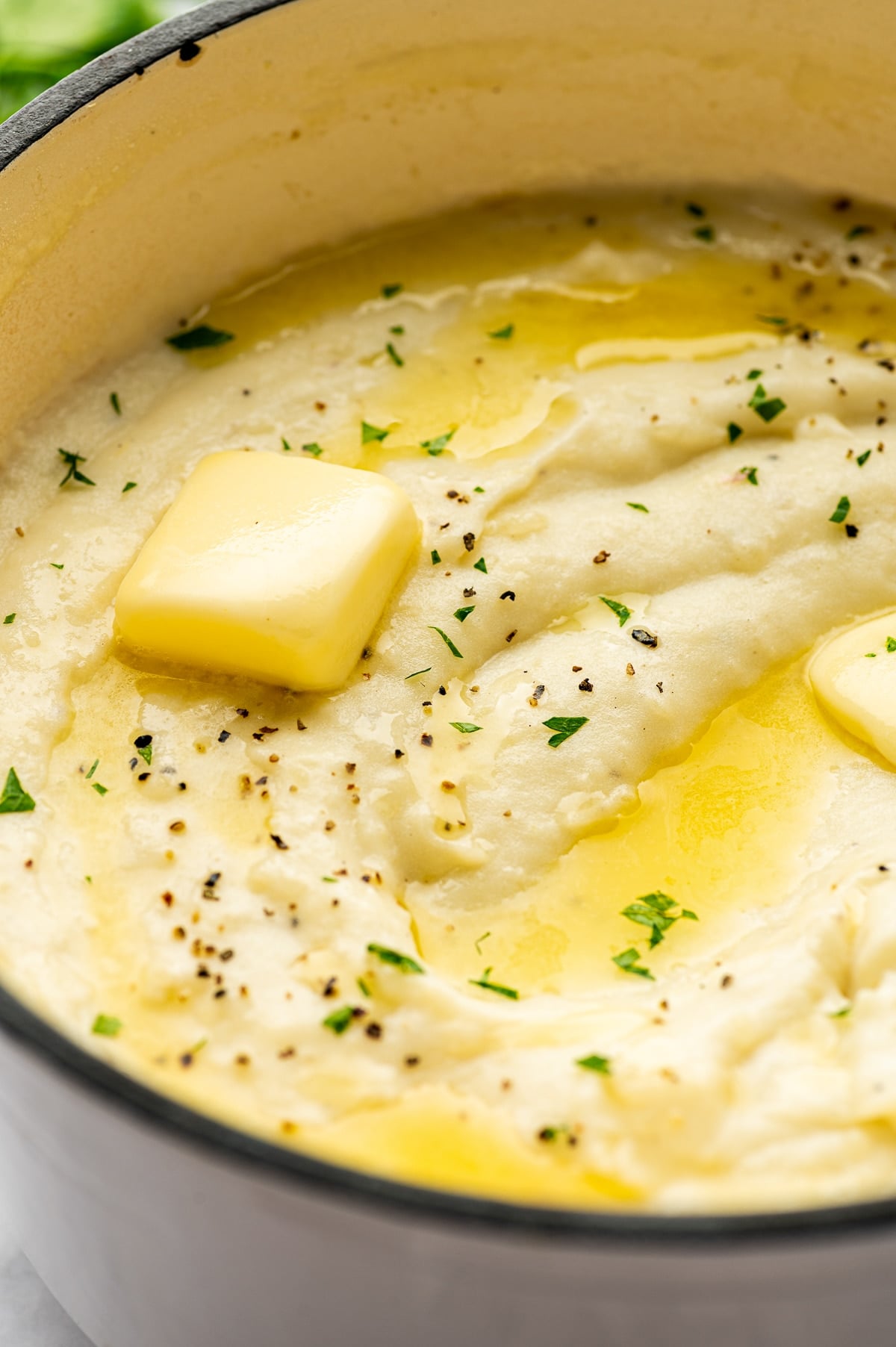 Close-up view of smooth, creamy mashed potatoes inside a cream-colored enameled pot, topped with a pat of melting pale yellow butter, pools of melted butter, chopped fresh green parsley, and cracked black pepper.