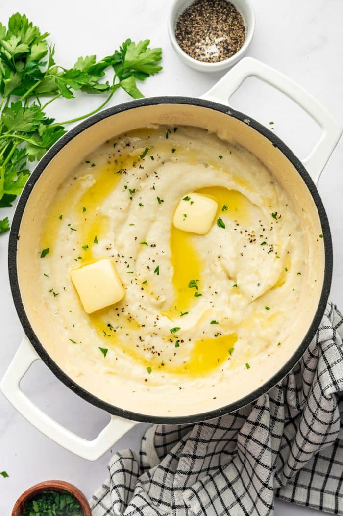 Overhead view of creamy mashed potatoes in a white enameled Dutch oven topped with two pats of pale yellow butter, melted butter, chopped fresh green parsley, and cracked black pepper. Fresh parsley, a small white bowl of black pepper, and a black-and-white checkered kitchen towel surround the pot on a light gray surface.