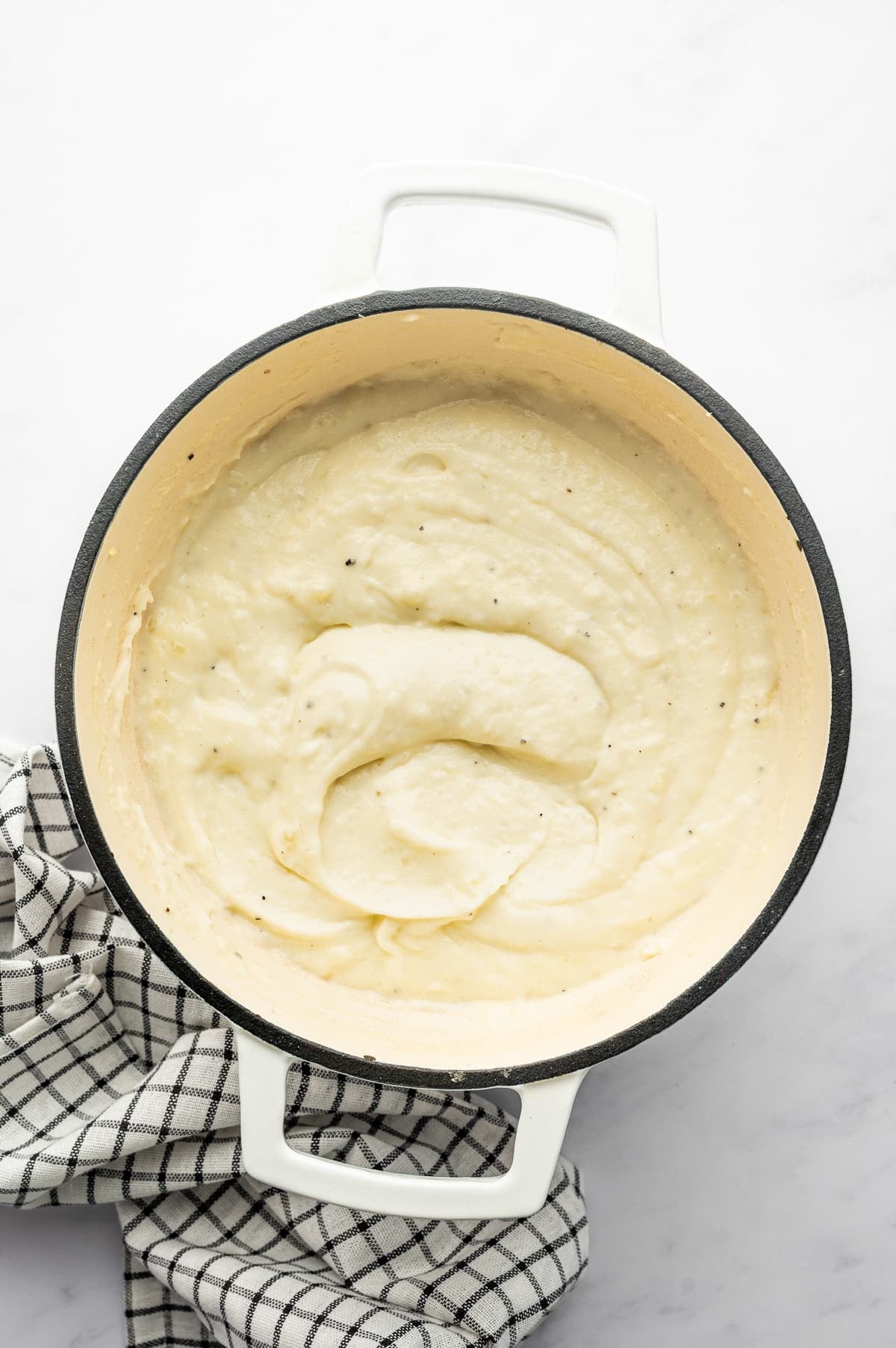 Overhead view of smooth, creamy mashed Russet potatoes in a white enameled Dutch oven with a cream-colored interior and dark rim. The potatoes have soft swirls and visible flecks of black pepper, with a black-and-white checkered kitchen towel beside the pot.