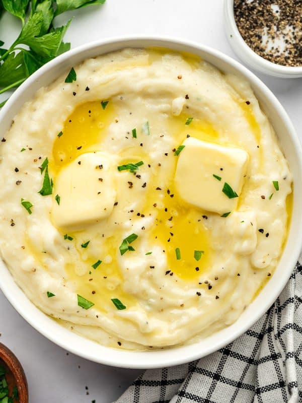 Overhead view of creamy mashed potatoes served in a large white bowl and topped with two pats of pale yellow butter, melted butter, chopped fresh parsley, and cracked black pepper. Fresh parsley, a small white bowl of black pepper, a small brown bowl of chopped parsley, and a black-and-white checkered kitchen towel surround the bowl on a light gray surface.