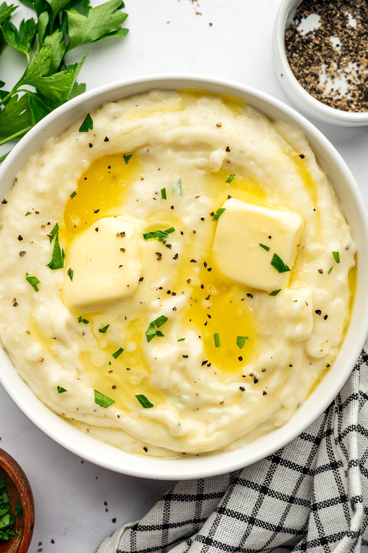 Overhead view of creamy mashed potatoes served in a large white bowl and topped with two pats of pale yellow butter, melted butter, chopped fresh parsley, and cracked black pepper. Fresh parsley, a small white bowl of black pepper, a small brown bowl of chopped parsley, and a black-and-white checkered kitchen towel surround the bowl on a light gray surface.