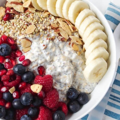 white bowl with sliced banana oats, over night yogurt, blueberries, raspberries, and blackberries