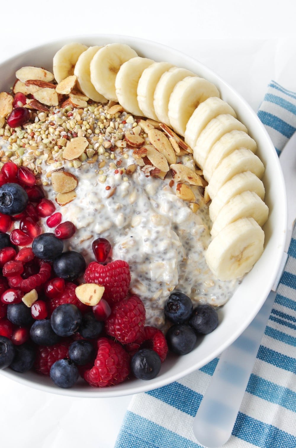 white bowl with sliced banana oats, over night yogurt, blueberries, raspberries, and blackberries
