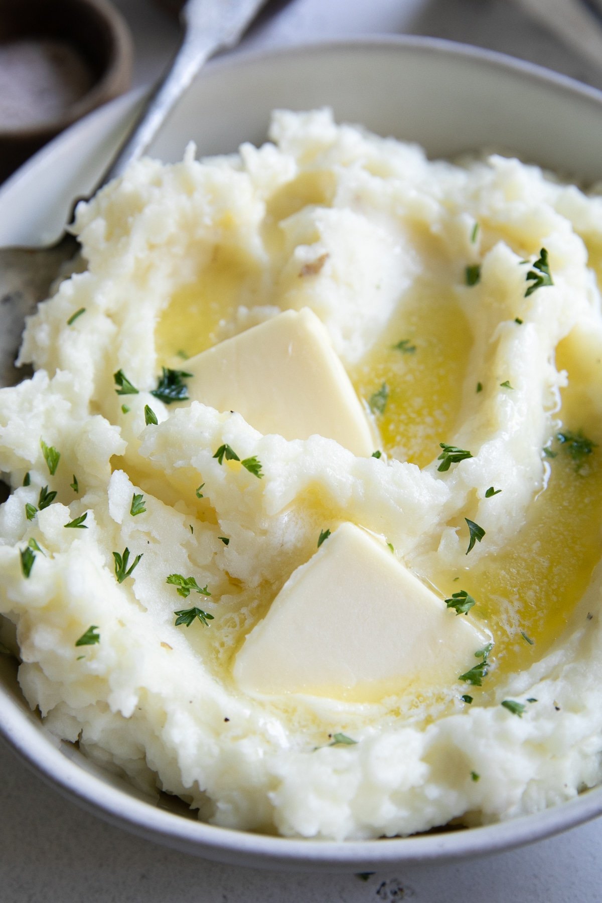 White shallow bowl filled with fluffy mashed potatoes, topped with two pats of butter, and garnished with fresh chopped parsley.