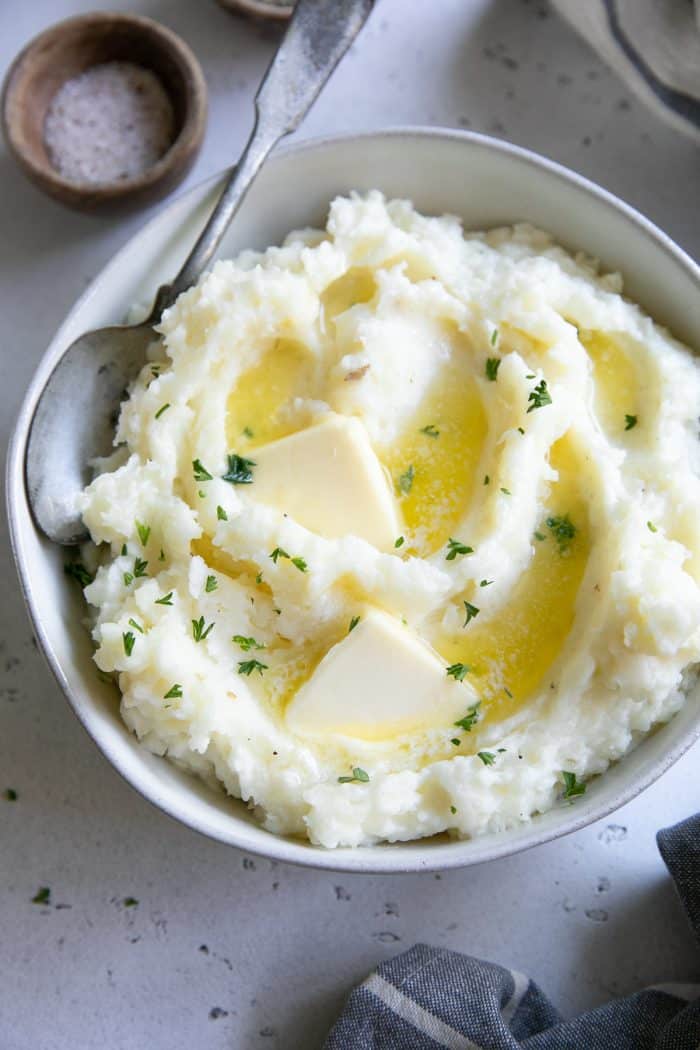 White shallow bowl filled with fluffy mashed potatoes, topped with two pats of butter, and garnished with fresh chopped parsley.