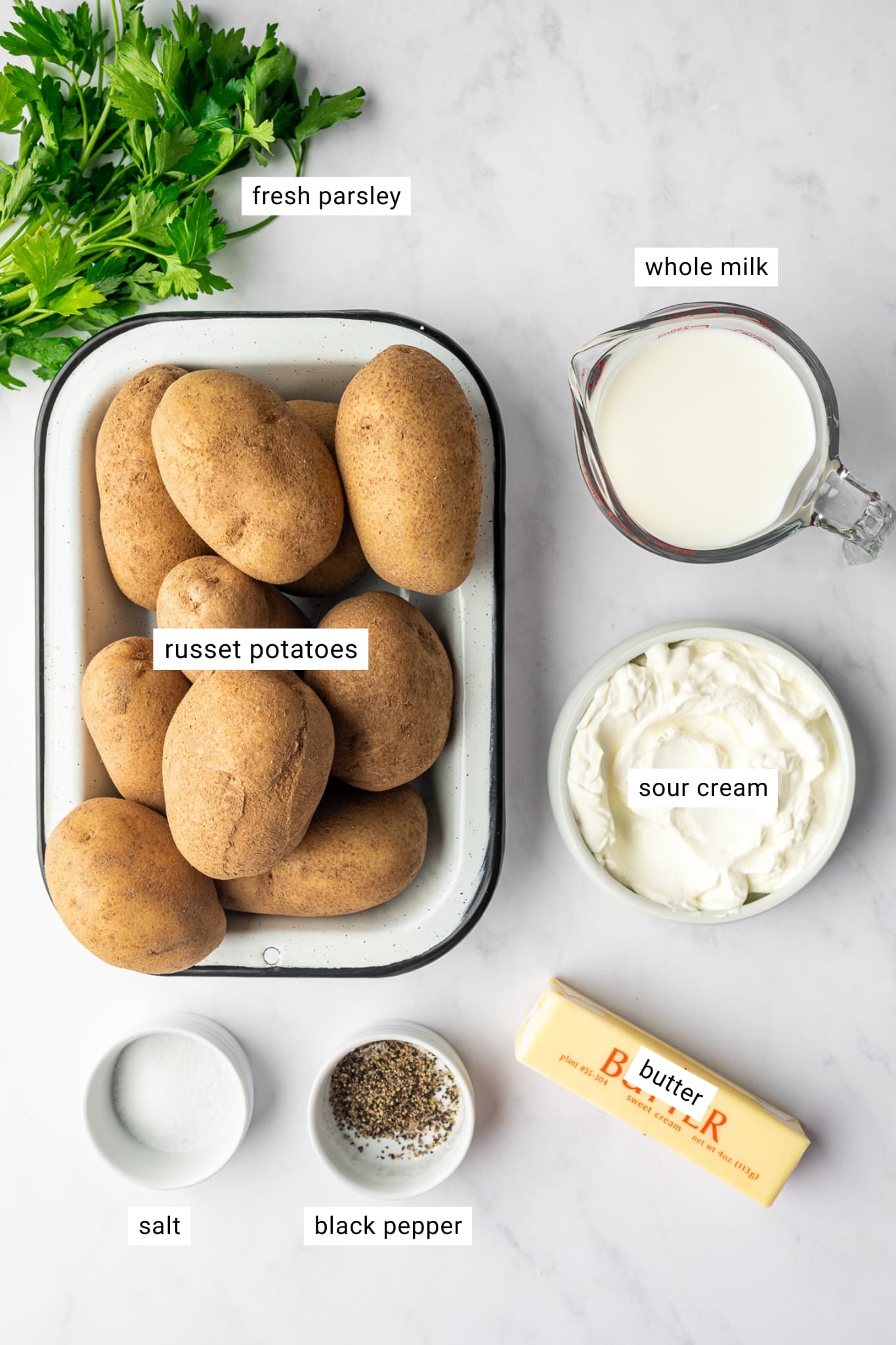 Overhead ingredient layout for creamy mashed potatoes showing whole Russet potatoes in a white enamel tray with a black rim, whole milk in a clear glass measuring cup, sour cream in a white bowl, a wrapped stick of butter, salt and black pepper in small white bowls, and fresh green parsley on a light gray surface. White text labels identify "fresh parsley," "whole milk," "russet potatoes," "sour cream," "salt," and "black pepper."