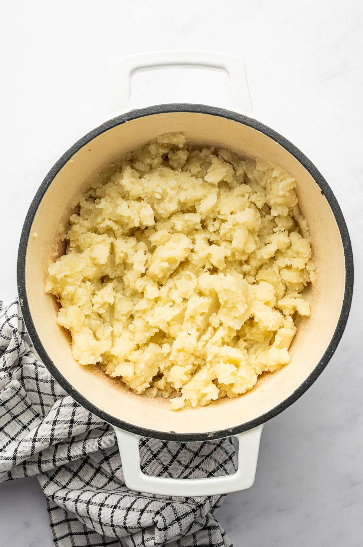 Overhead view of roughly mashed Russet potatoes in a white enameled Dutch oven with a cream interior and dark rim. The potatoes are dry and lightly textured before the warm dairy mixture is added, with a black-and-white checkered kitchen towel beside the pot. No text overlay or garnish is shown.