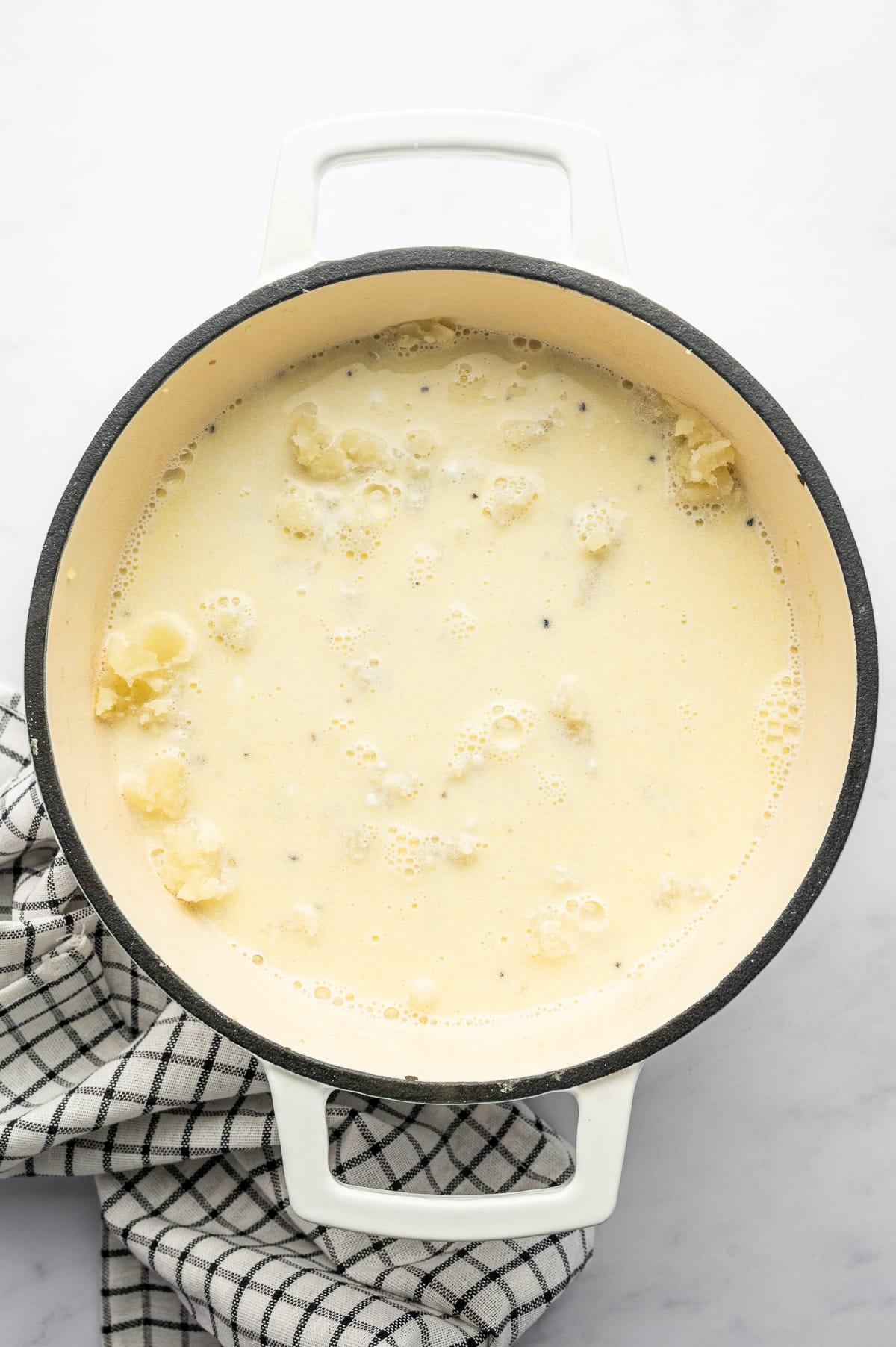 Overhead view of warm creamy dairy mixture poured over partially mashed Russet potatoes in a white enameled Dutch oven with a cream interior and dark rim. Small pieces of potato are visible beneath the pale buttery liquid, with a black-and-white checkered kitchen towel beside the pot.