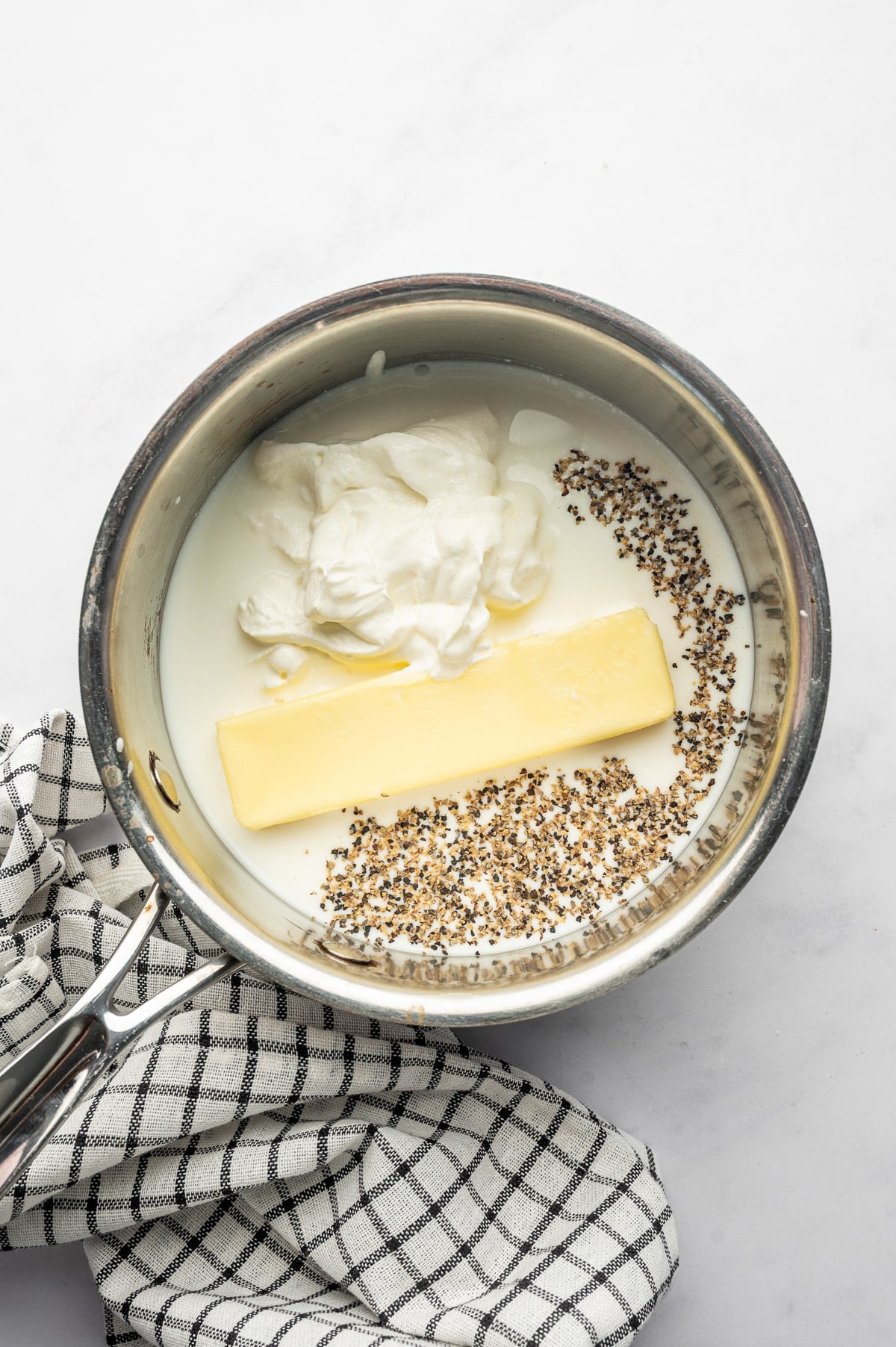 Overhead view of whole milk, a stick of pale yellow butter, dollops of white sour cream, and cracked black pepper in a small stainless steel saucepan on a light gray surface. A black-and-white checkered kitchen towel rests beside the pan. No text overlay or garnish is shown.
