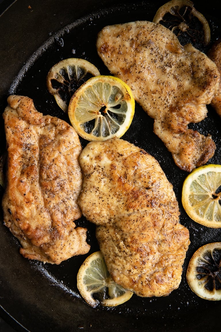 Cooked chicken cutlets in a large cast iron skillet with lemon slices.