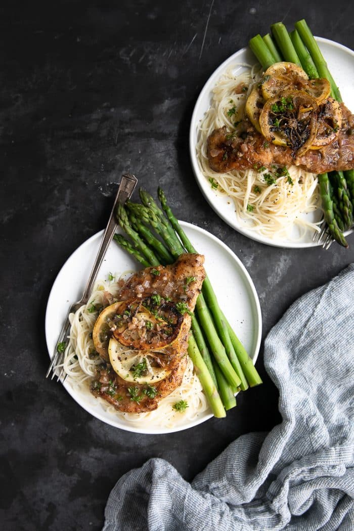 Two white plates with angel hair pasta, asparagus, and fully prepared chicken piccata cutlet with piccata sauce and capers.