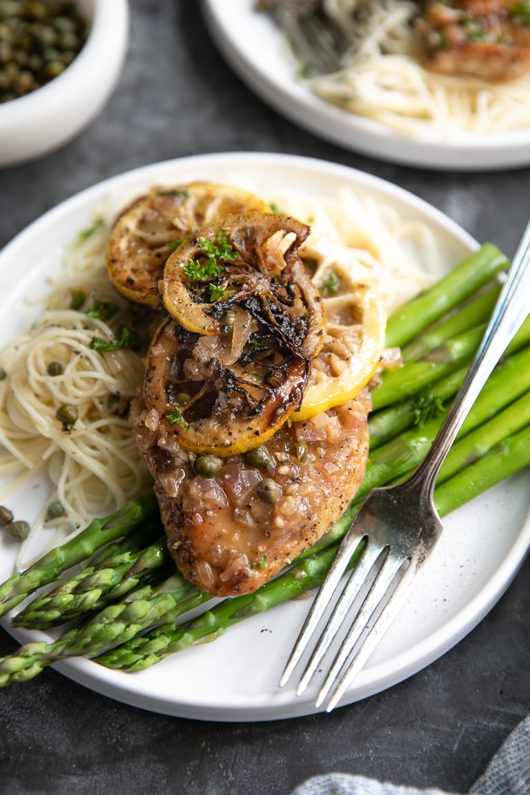 White plate with cooked noodles, asparagus, and one chicken piccata cutlet topped with lemon piccata sauce.