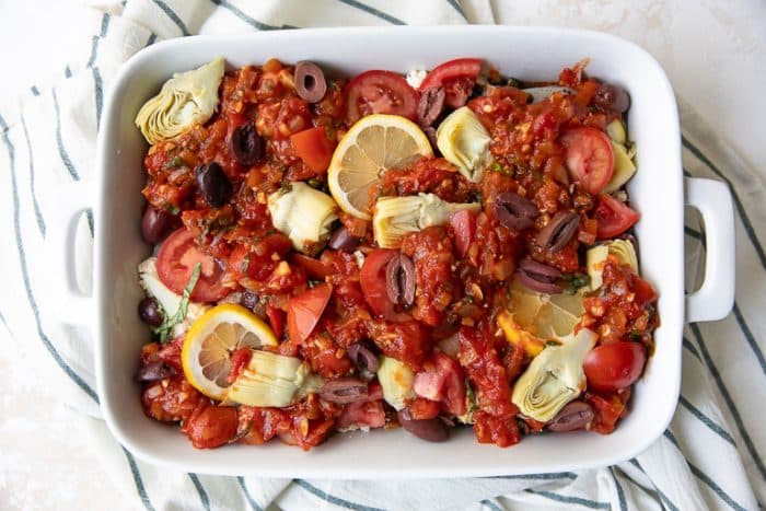 a layer of artichokes and tomatoes being added to Mediterranean baked fish in a casserole dish