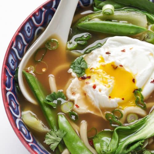 Blue bowl filled with udon noodle soup made with bok choy, snap peas, green onions, and a poached egg.