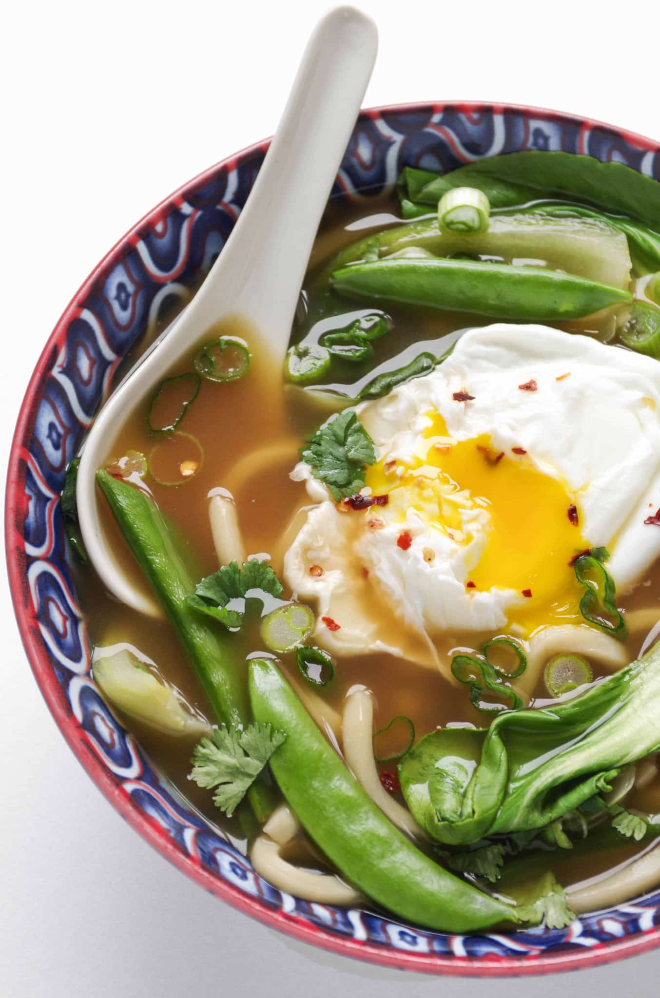 Blue bowl filled with udon noodle soup made with bok choy, snap peas, green onions, and a poached egg.