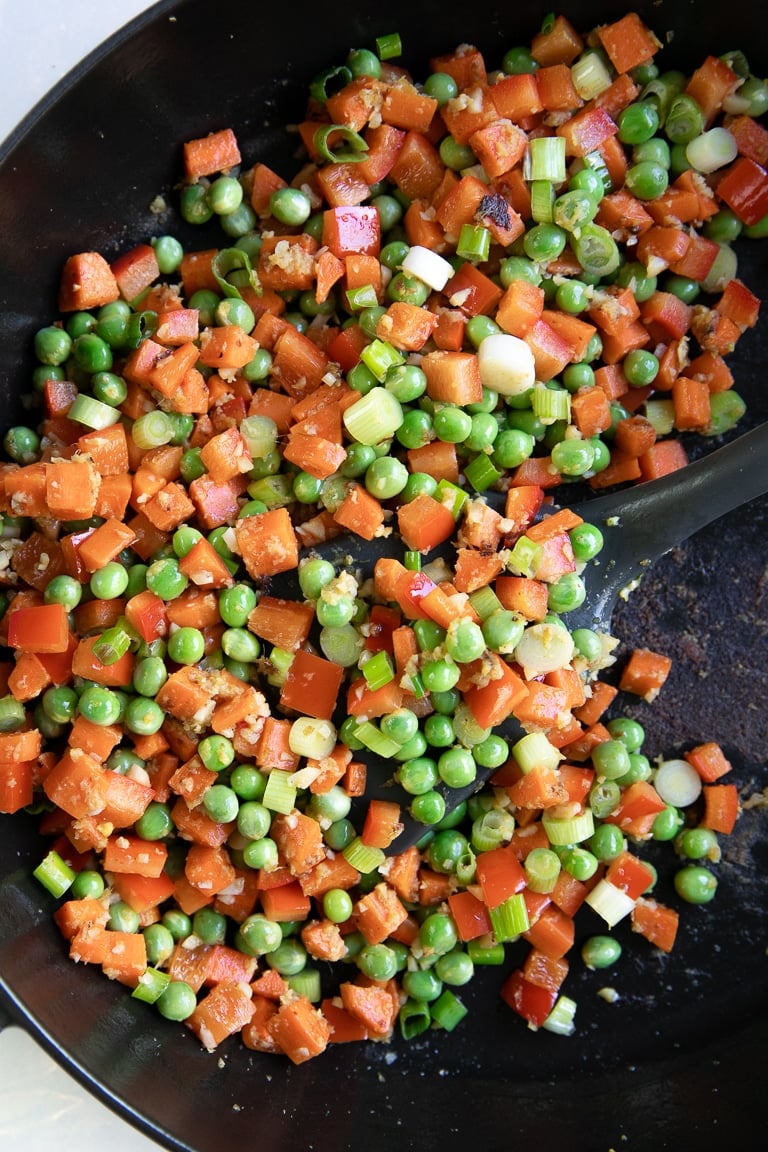 Diced carrot and red bell pepper with peas cooking in a large heavy-bottomed skillet.