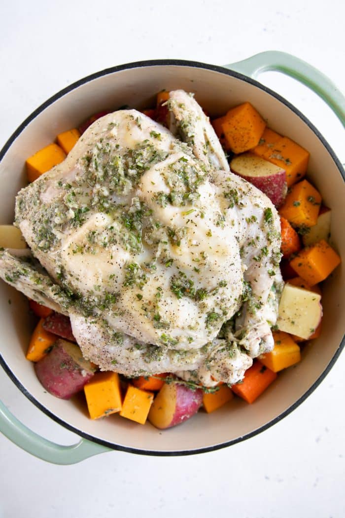 Overhead image of a large Dutch oven filled with chopped potatoes, carrots, and butternut squash topped with a whole raw chicken covered in a garlic herb and butter sauce.