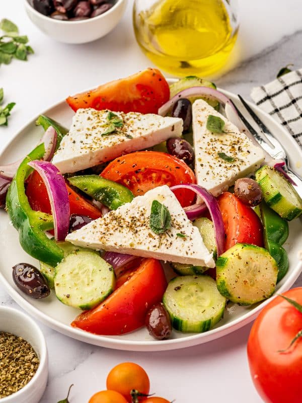 Close-up of classic Greek salad on a light beige plate with tomato wedges, cucumber rounds, red onion, green bell pepper, Kalamata olives, and large triangular pieces of feta topped with dried oregano, with a glass bottle of olive oil, extra olives, oregano, and tomatoes nearby.