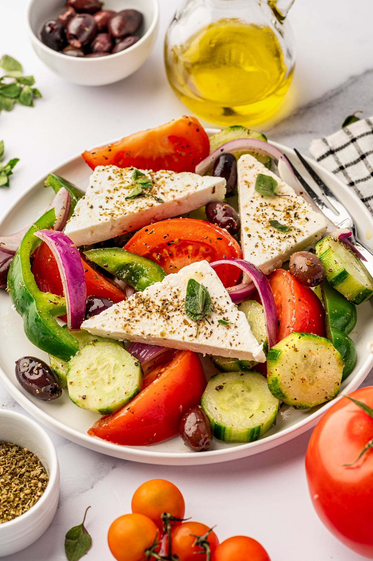 Close-up of classic Greek salad on a light beige plate with tomato wedges, cucumber rounds, red onion, green bell pepper, Kalamata olives, and large triangular pieces of feta topped with dried oregano, with a glass bottle of olive oil, extra olives, oregano, and tomatoes nearby.