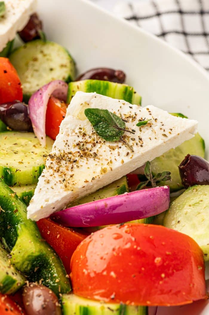 Close-up of a triangular slab of feta cheese topped with dried oregano and fresh oregano on a Greek salad with tomato wedges, cucumber rounds, red onion, green bell pepper, and Kalamata olives.