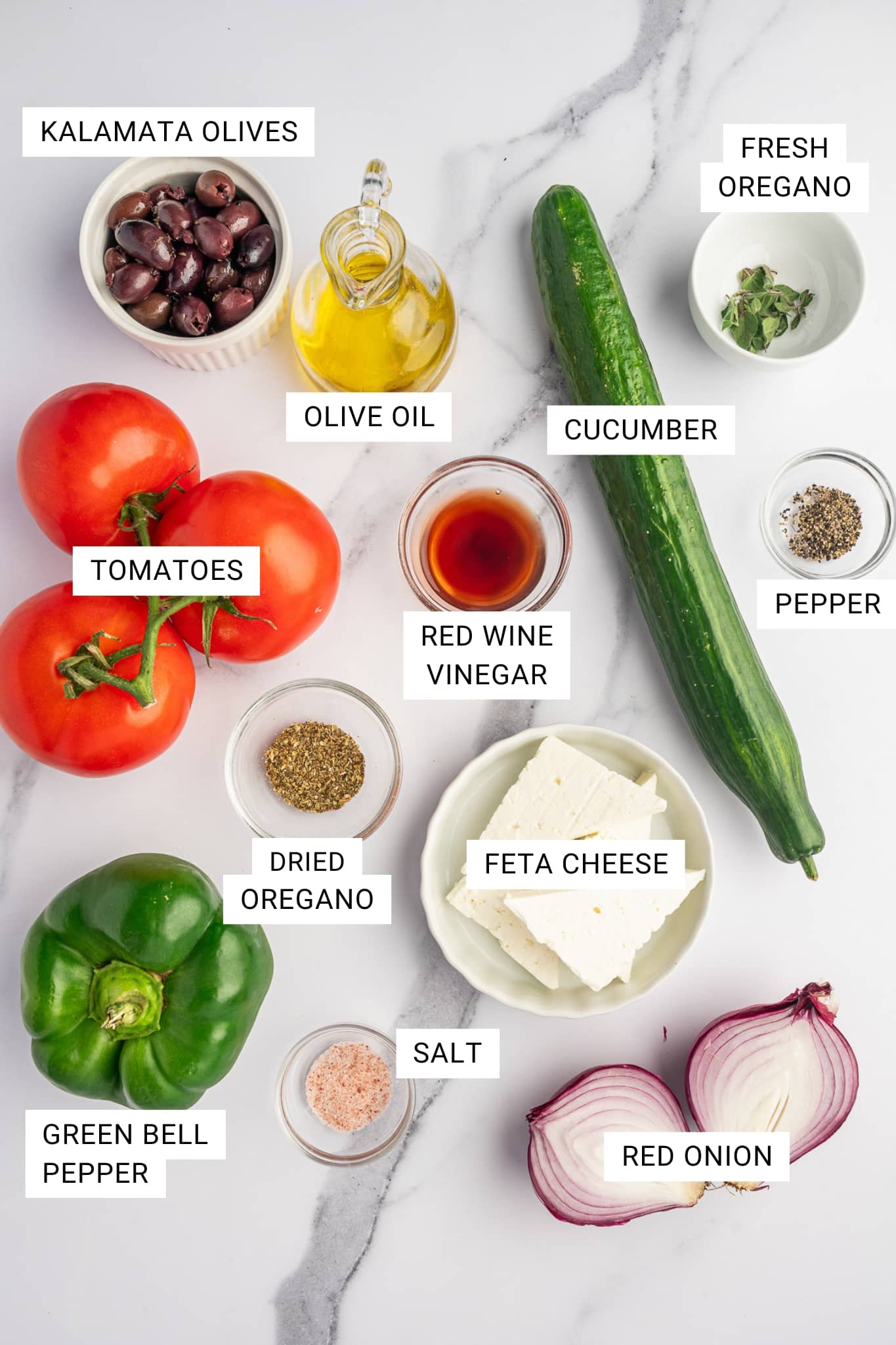 Overhead labeled ingredient photo for Greek salad on a white marble surface with text overlays for Kalamata olives, olive oil, fresh oregano, cucumber, pepper, tomatoes, red wine vinegar, feta cheese, dried oregano, salt, green bell pepper, and red onion.