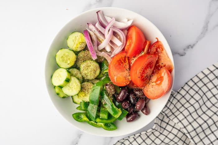 Overhead view of Greek salad ingredients in a white bowl, including sliced cucumber, tomato wedges, red onion, green bell pepper, Kalamata olives, dried oregano, salt, and black pepper, with a black-and-white checkered towel on the side.