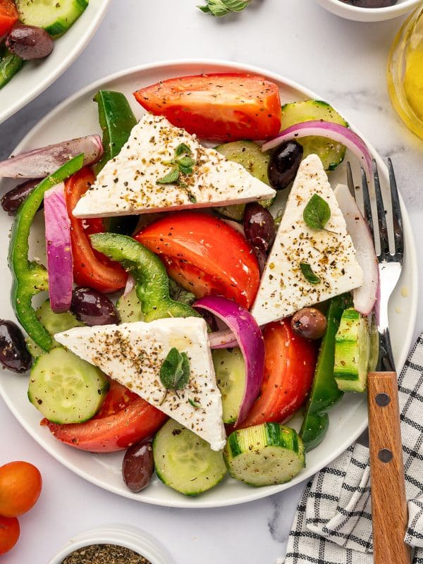 Close-up overhead view of traditional Greek salad with tomato wedges, cucumber rounds, red onion, green bell pepper, Kalamata olives, and triangular feta slabs sprinkled with dried oregano, with a wooden-handled fork and black-and-white checkered towel on the side.